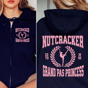 May include: Navy blue zip-up hoodie with the text "NUTCRACKER GRAND PAS PRINCESS" in pink. The design includes a ballerina silhouette within a laurel wreath. The hoodie has a drawstring hood and a front zipper.