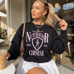 May include: A black crewneck sweatshirt featuring a pink graphic print that reads Nutcracker Chinese, Established 1892, and Classical Ballet, with a central shield emblem containing a silhouette of a ballerina in a tutu.