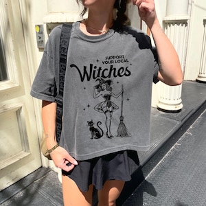 May include: A charcoal gray short-sleeve t-shirt featuring a black graphic print of a witch, a cat, and a broomstick. The text reads Support Your Local Witches, accompanied by small star icons. The shirt has a crew neckline and a relaxed fit.