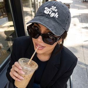 May include: A dark gray baseball cap with the text "Be Sigh" in white script. The cap is worn with black sunglasses and a black blazer. A clear plastic cup of iced coffee with a straw is held in the hand.