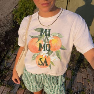 Può includere: T-shirt bianco con grafica "Mimosa" con arance e fiori. La maglietta è abbinata a pantaloncini a fantasia verdi e bianchi. Sono visibili anche una collana d'oro e una borsa tote beige.