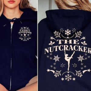 Puede incluir: Sudadera con capucha azul marino con cremallera y un gráfico blanco de una bailarina y el texto "THE NUTCRACKER" rodeado de copos de nieve. La sudadera tiene una capucha con cordón.