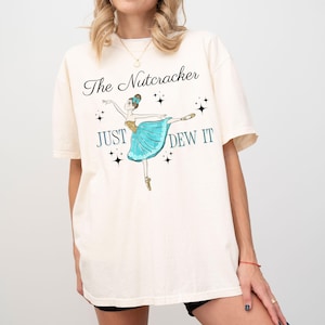 May include: A white t-shirt with a graphic of a ballerina in a blue tutu. The text "The Nutcracker Just Dew It" is printed on the shirt.