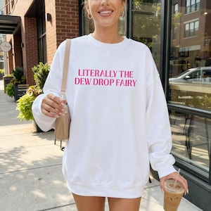 May include: A white long-sleeve crewneck sweatshirt featuring the text LITERALLY THE DEW DROP FAIRY printed in bold pink capital letters across the chest. The garment has a relaxed fit with ribbed cuffs and a ribbed neckline.
