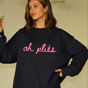 May include: Black sweatshirt with the phrase "oh pliés" in pink script. The sweatshirt is a crew neck style and appears to be made of a soft material. The overall aesthetic is casual and comfortable.