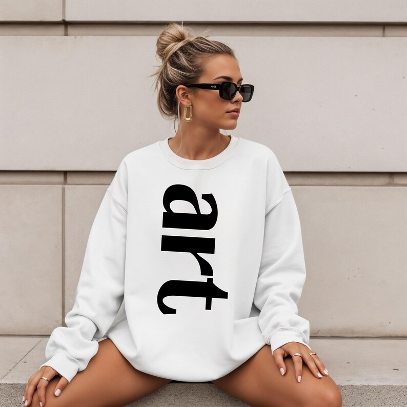May include: White sweatshirt with the word "art" printed vertically in large, black letters. The sweatshirt has long sleeves and a crew neck. The overall style is casual and minimalist.