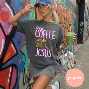 May include: Dark gray t-shirt with the text "iced COFFEE & JESUS" in pink and orange. The shirt features a graphic of a coffee cup and a bible. The person is wearing a blue cap and holding a green drink.
