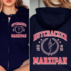 May include: Navy blue zip-up hoodie with a Nutcracker and Marzipan design in pink and white. The design includes a ballerina, stars, and the text "ES TD 18 92".