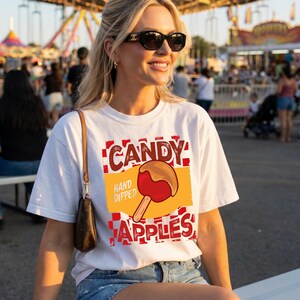 May include: A white short-sleeve t-shirt featuring a graphic of a red candy apple with the text CANDY APPLES and HAND DIPPED. The design includes a red and white checkered pattern background and a yellow rectangular accent behind the apple illustration.