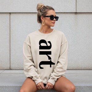 May include: A beige sweatshirt with the word "art" printed vertically in large, black letters. The sweatshirt is a neutral color and has a crew neck. The image shows a person wearing the sweatshirt.