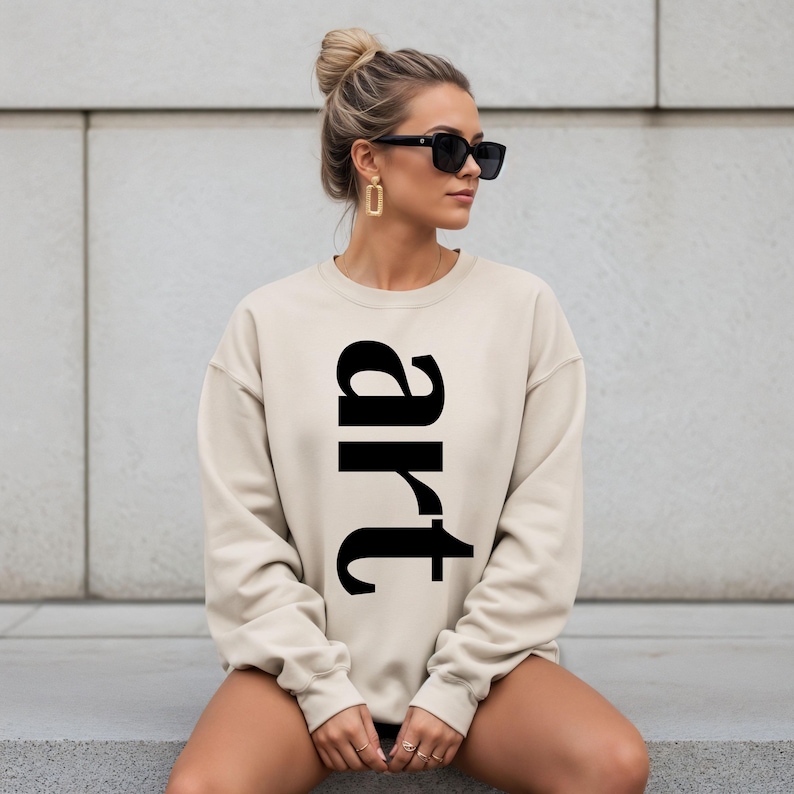May include: A beige sweatshirt with the word "art" printed vertically in large, black letters. The sweatshirt is a neutral color and has a crew neck. The image shows a person wearing the sweatshirt.