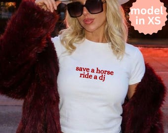 Puede incluir: Top corto blanco con el texto rojo "save a horse ride a dj". La modelo lleva un sombrero de vaquero blanco, gafas de sol grandes y un abrigo de piel sintética burdeos. La modelo lleva una talla XS.