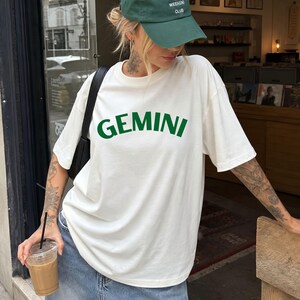 May include: A white short-sleeved t-shirt featuring the word GEMINI printed in a bold, dark green sans-serif font across the chest. The shirt has a crew neckline and a loose, relaxed fit, paired with blue denim jeans and a dark green baseball cap.