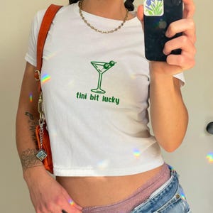 Op de afbeelding: Wit T-shirt met een groene martini-glas afbeelding en de tekst "tini bit lucky".