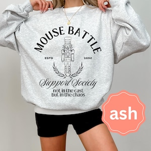 May include: A light grey sweatshirt with a black graphic that reads "MOUSE BATTLE" above a nutcracker illustration. Below the graphic is "Support Society" and the text "not in the cast but in the chaos". The sweatshirt has a crew neck.