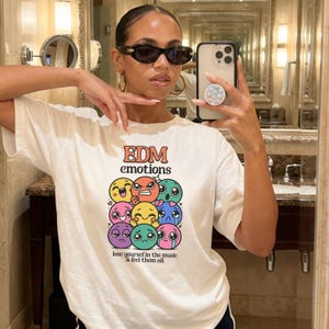 May include: White t-shirt with the text "EDM emotions" in orange and a graphic of colorful cartoon faces expressing different emotions. The phrase "lose yourself in the music & feel them all" is below the graphic.