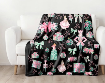 Puede incluir: Una manta de forro polar negra con un estampado navideño festivo que presenta un cascanueces, una bailarina, regalos, adornos y lazos. El estampado es en tonos de rosa, verde y blanco sobre un fondo negro.