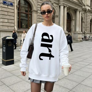 May include: A white sweatshirt with the word "art" printed vertically in large, black letters. The sweatshirt is worn by a person in an outdoor setting, paired with a gray skirt and sunglasses. The person is holding a coffee cup.