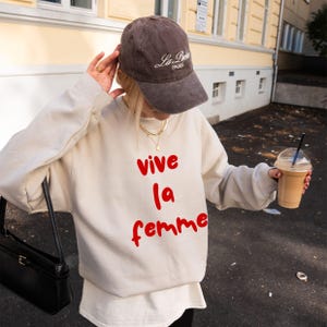 Może przedstawiać: Osoba nosząca brązową czapkę z daszkiem z napisem "La Paris" i kremową bluzę z napisem "Vive la femme" w kolorze czerwonym. Osoba trzyma czarną torebkę i napój kawowy.