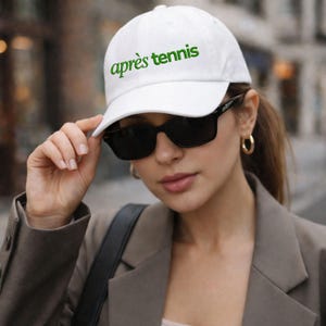 May include: White baseball cap with the text "après tennis" in green. The cap is worn by a person wearing black sunglasses and a brown blazer. The background is blurred, suggesting an outdoor setting.