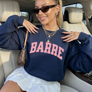 May include: Navy blue sweatshirt with the word "BARRE" in pink block letters. The sweatshirt is paired with a white pleated skirt. The person is wearing sunglasses and gold jewelry.