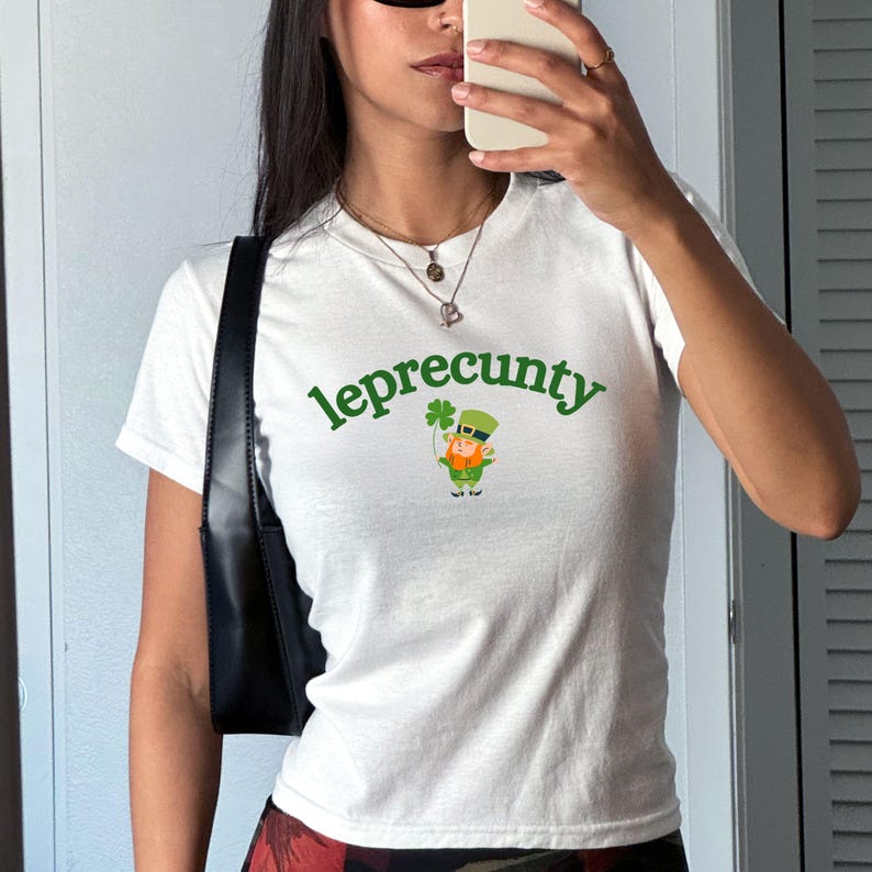 May include: White t-shirt with the word "leprecunty" in green, arched above a cartoon leprechaun holding a shamrock. The leprechaun wears a green hat and orange beard. The shirt is short-sleeved.