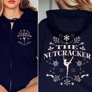 May include: A navy blue zip-up hoodie with a white design on the front and back. The front design says "The Nutcracker" with a ballerina silhouette. The back design says "The Nutcracker" with a ballerina silhouette and a snowflake pattern.