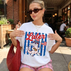 以下が含まれることがあります： 愛国的なデザインの白いTシャツ。「Later」の文字が赤い筆記体で描かれ、アンクルサムの帽子とサングラスをかけ、ホットドッグとアメリカ国旗を持った漫画のアリゲーターが描かれています。