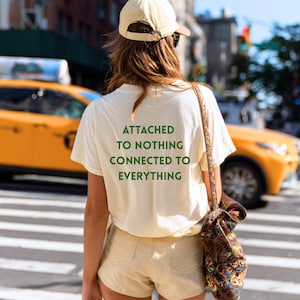 May include: Cream-colored t-shirt with the text "ATTACHED TO NOTHING CONNECTED TO EVERYTHING" in green. The person is wearing tan shorts and a baseball cap. A patterned shoulder bag is visible. A yellow taxi is in the background.