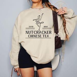 Puede incluir: Sudadera color crema con un gráfico negro de una bailarina y el texto "NUTCRACKER CHINESE TEA" con "ESTD 1892" arriba y abajo. La sudadera se combina con pantalones cortos negros.