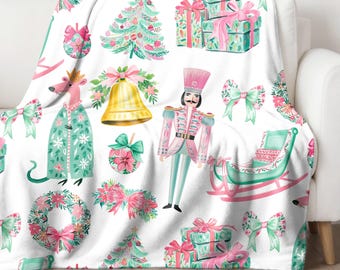 Puede incluir: Una manta blanca con un patrón navideño festivo que incluye un cascanueces, un trineo, un ratón, una campana, una corona y un árbol de Navidad. El patrón está en tonos de rosa, verde y blanco.