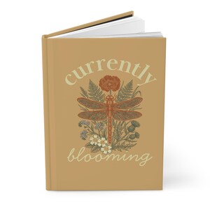 May include: A tan journal with the words "currently blooming" in a script font. The cover features a detailed illustration of a dragonfly, flowers, and foliage in shades of brown and cream. The journal is open to reveal white pages.