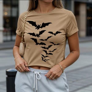 May include: Tan crop top featuring a black bat graphic. The bats are arranged in a descending flight pattern. The shirt has short sleeves and a crew neck. The overall theme is Halloween.