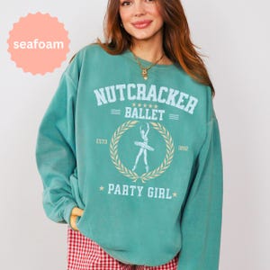 May include: A seafoam green crewneck sweatshirt featuring a graphic print with the text NUTCRACKER BALLET PARTY GIRL, ESTD 1892, and a central illustration of a ballerina inside a laurel wreath. The design includes five stars above the ballerina.