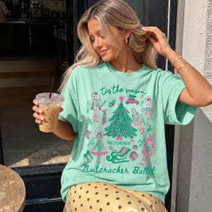 May include: A light green t-shirt featuring a graphic print with the text 'Tis the season' and 'Nutcracker Ballet'. The design includes a central Christmas tree surrounded by illustrations of a nutcracker, a ballerina, a sleigh, and gifts in pink and green ink.