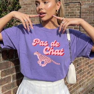 Op de afbeelding: Een paars T-shirt met de tekst "Pas de Chat" in roze en rood, en een roze luipaardafbeelding. Het shirt is gemaakt van een zacht materiaal en is geschikt voor vrijetijdskleding.