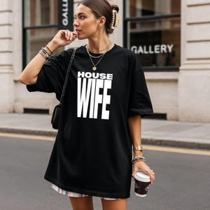 Könnte beinhalten: Schwarzes übergroßes T-Shirt mit dem Wort "HOUSEWIFE" in fetten weißen Buchstaben aufgedruckt. Das Shirt wird von einer Person getragen, und das Bild wird im Freien aufgenommen. Die Person trägt auch Schmuck und hält eine Kaffeetasse.