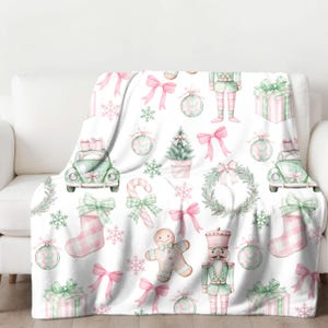 Puede incluir: Una manta de forro polar blanca con un patrón repetitivo de ilustraciones navideñas de color rosa y verde. Las ilustraciones incluyen un hombre de jengibre, un cascanueces, un árbol de Navidad, una corona, un bastón de caramelo, una media, un coche con regalos y un lazo.