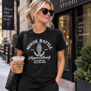 May include: A black short-sleeved t-shirt featuring a white graphic of a nutcracker with the text MOUSE BATTLE ESTD 1892 Support Society not in the cast but in the chaos. The shirt has a crew neckline and a standard fit, made of cotton fabric.