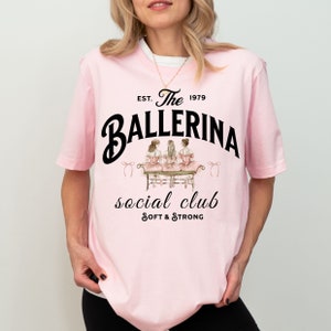 May include: Pink t-shirt with a graphic of three ballerinas sitting on a bench. The text "The Ballerina Social Club Est. 1979 Soft & Strong" is printed on the shirt.