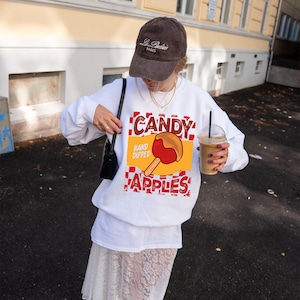 May include: A white sweatshirt with a graphic of a candy apple, the words "CANDY HAND DIPPED APPLES" in red and brown. The person is wearing a brown baseball cap and holding a coffee drink.