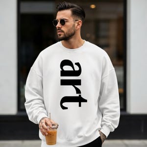 May include: White sweatshirt with the word "art" printed vertically in large, black letters. The sweatshirt is a casual style, suitable for everyday wear. The image shows a person wearing the sweatshirt.