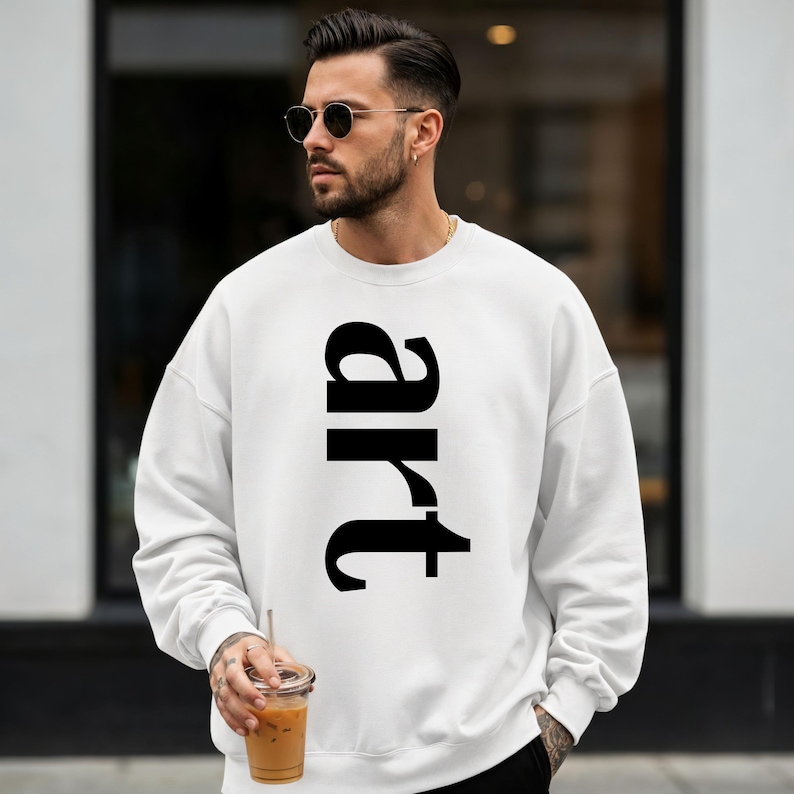 May include: White sweatshirt with the word "art" printed vertically in large, black letters. The sweatshirt is a casual style, suitable for everyday wear. The image shows a person wearing the sweatshirt.
