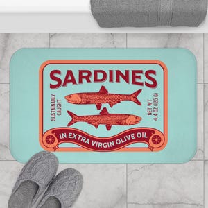May include: A rectangular bath mat with a light blue background and a sardine can design. The design features two red sardines, the word "SARDINES", and the text "IN EXTRA VIRGIN OLIVE OIL". The net weight is 4.4 oz (125 g).