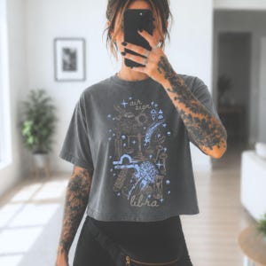 May include: A charcoal grey cropped t-shirt featuring a light blue and white graphic print with the text "air sign" and "libra". The design includes celestial and astrological symbols such as scales, stars, a sun, a moon, and an hourglass.