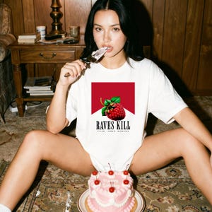May include: White t-shirt with a graphic design featuring a raspberry and the text "RAVES KILL". A pink cake with cherries sits in front of the shirt. The shirt's design is set against a red background.