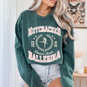 May include: A green long-sleeved sweatshirt with a pink graphic print that says "Nutcracker Classical Season Ballerina" with a ballerina silhouette and the text "Est. 1892".