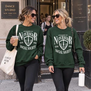 May include: A dark green crewneck sweatshirt featuring a white graphic print that reads "ESTABLISHED 1892 NUTCRACKER SOCIAL SOCIETY" with a central shield emblem containing a nutcracker figure, flanked by the words "CAFFEINE" and "GASOLINE".