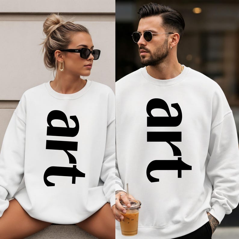 May include: White sweatshirts with the word "art" printed vertically in large, black letters. The sweatshirts are worn by two people, one with sunglasses and the other with a drink.