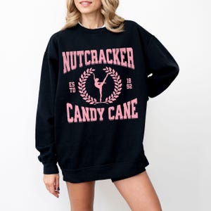 May include: Black sweatshirt with pink text that reads "NUTCRACKER CANDY CANE" with a ballerina graphic. The sweatshirt also has the text "ESTD 18 92". The sweatshirt is a crew neck style.